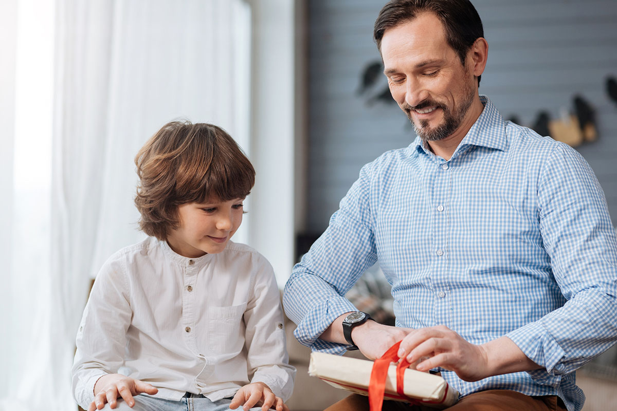 Lets look. Delighted handsome bearded man holding a present and opening it while sitting together with his son