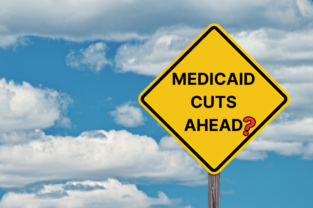 Image of street sign in Rhode Island with yellow sign that says Medicaid Cuts Ahead? in black lettering with a red question mark. Behind the sign, the clear East Greenwich sky shines brightly.